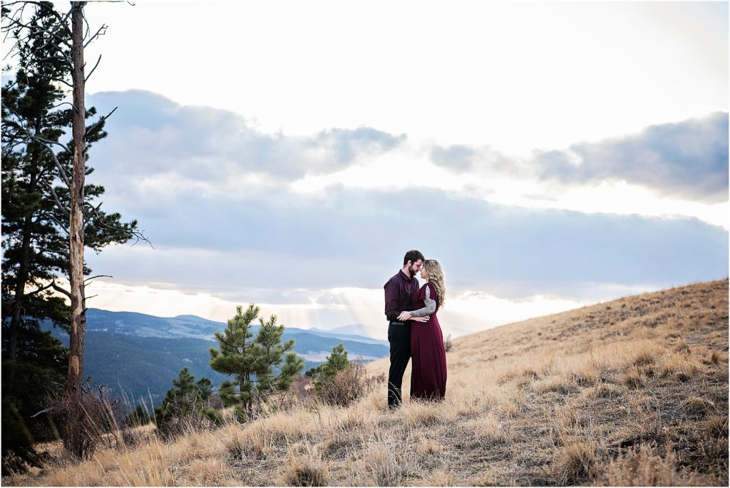 Andrew and Martha stand embracing in Colorado Rocky Mountains as the shines through the clouds close to sunset