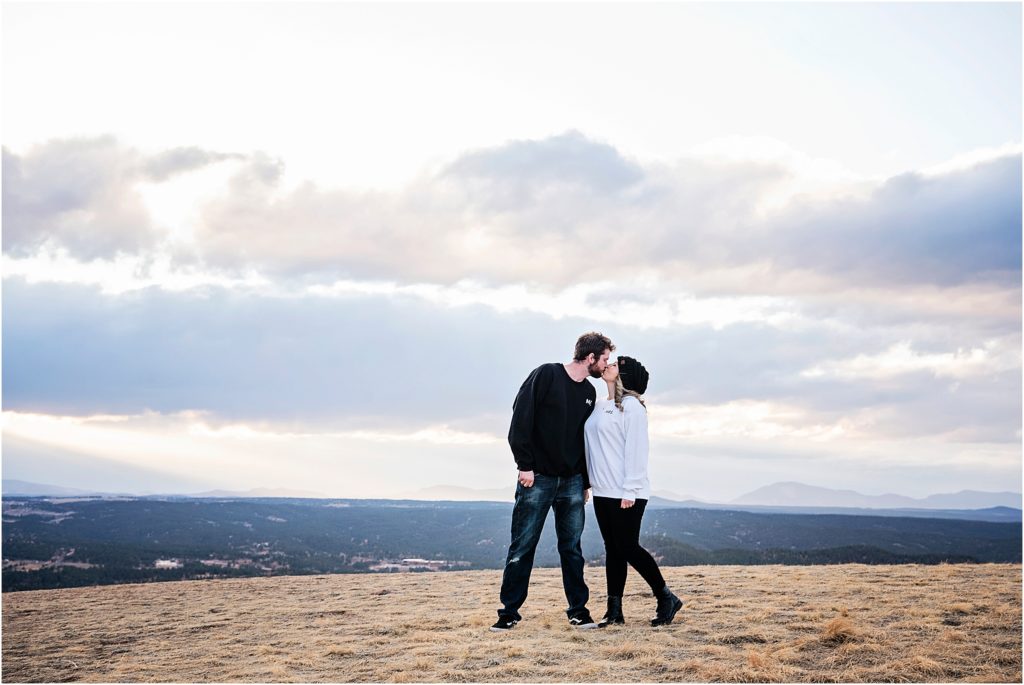 Couple stand and kiss on top of a mountain with the sunset filtered by blue and pink clouds.