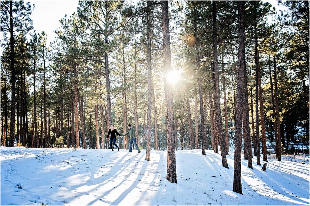couple walks through the snow in a forest with the sunlight filtered through the trees.