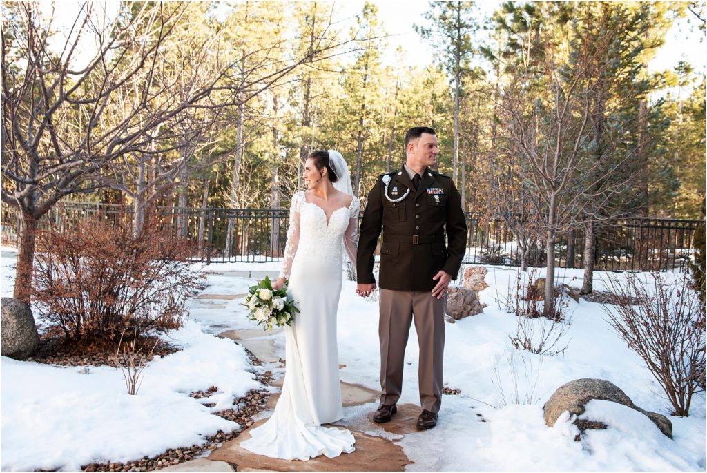 Bride and groom in uniform stand holding hands on a stone path with trees behind and snow all around