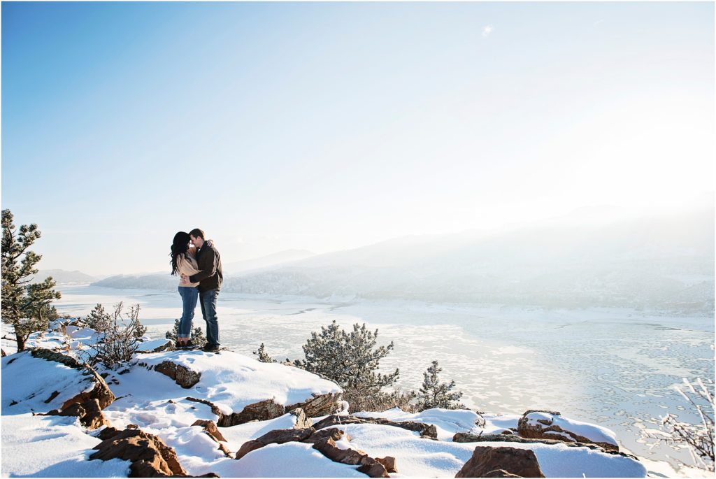 Couple stands together embracing with gorgeous sunset and mountain views behind them at Horsetooth Reservoir in Colorado