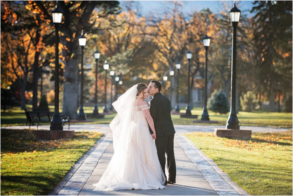 A newlywed groom kisses his brides temple as they walk down a long fall leaf covered pathway lined with lit lampposts at sunset at Colorado College