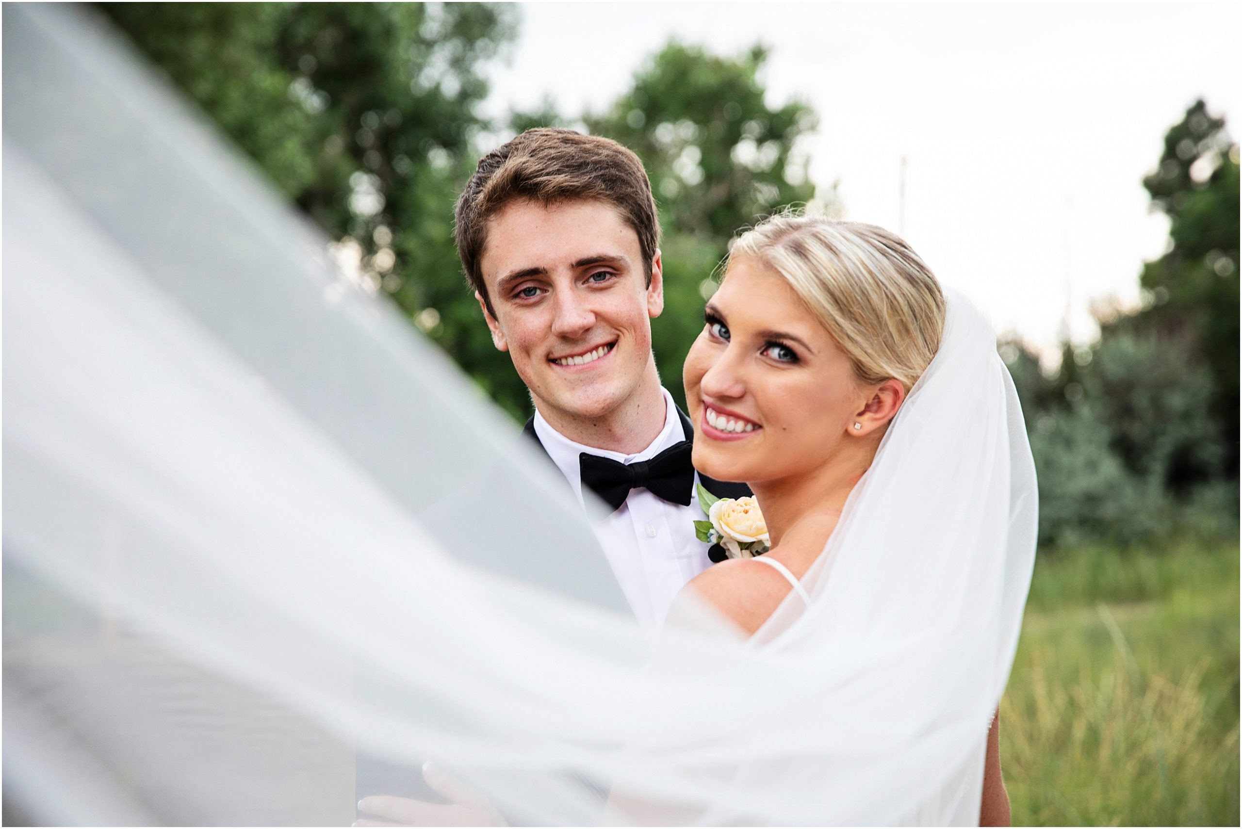 Bride looks back as her long veil sweeps fancifully over her shoulder her groom in his black bow tie smiles on