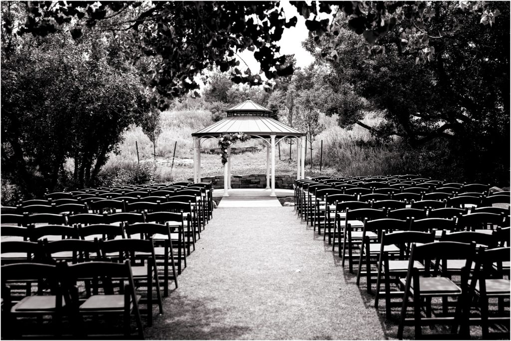 Outdoor ceremony surrounded by trees and greenery aisle leads to outdoor gazebo with simple floral arrangement black folding chairs complete the simple outdoor ceremony