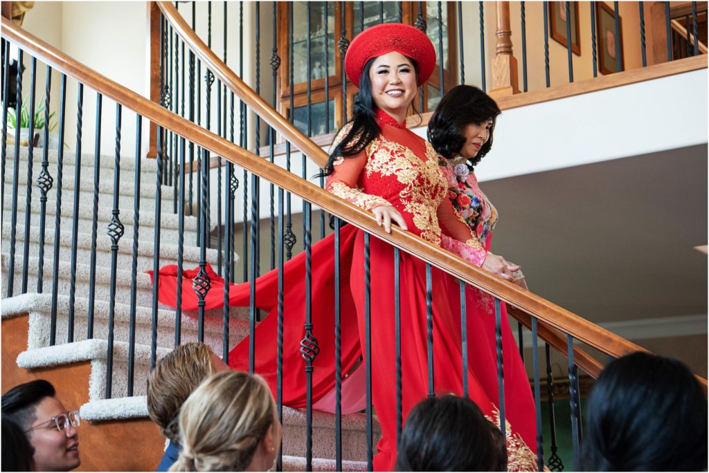 Bride glows in her red and gold tea dress as she descends the stairs with her mother