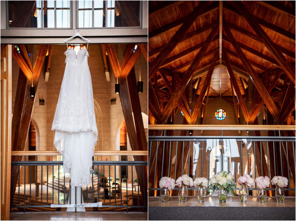 Stunning photograph of lace wedding dress hanging from cedar wood beams; a stained glass window is visible in the church behind the pink and white peony bouquets