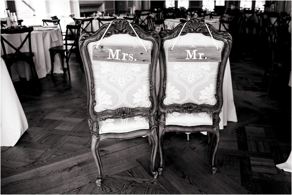 Bride and groom table and chairs with simple rustic signs hanging from the back