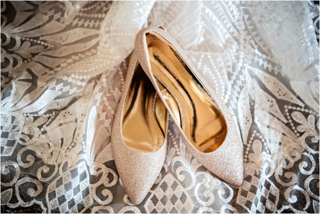 Rose gold sparkly wedding shoes are displayed atop unique lace pattern on wedding dress
