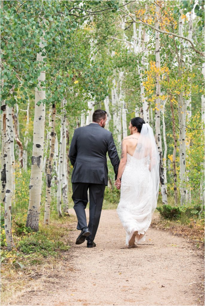 Romantic wedding photo of bride and groom hand-in-hand as they walk down a forested path surrounded by Aspen trees in outdoor Colorado wedding