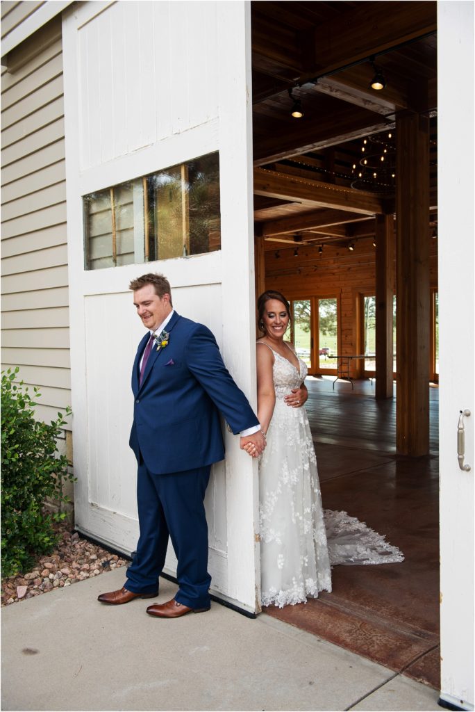 Bride and groom stand on either side of a white barn door holding hands as part of their first look photo