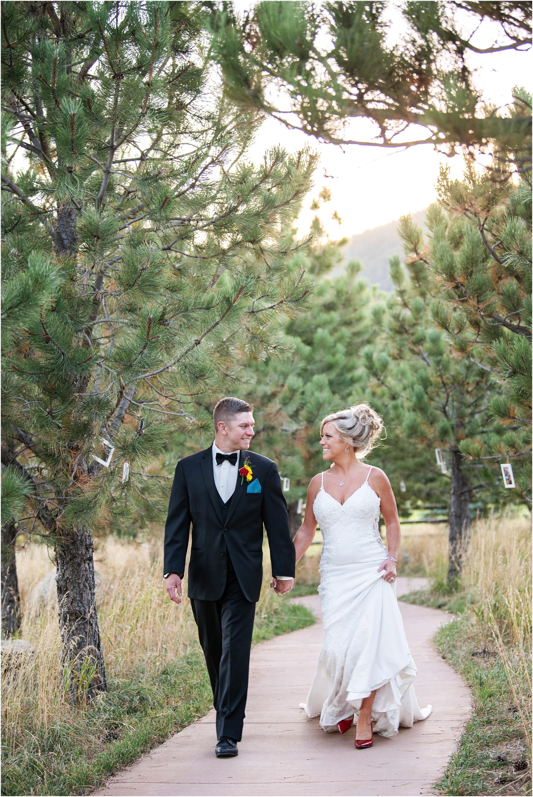 Bride and groom walk and hold hands down a cement path through trees smiling at each other