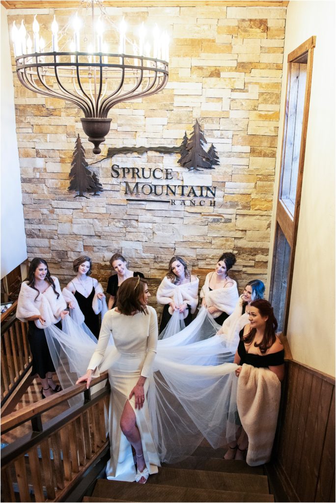 Bride stands on stairs at Spruce Mountain Ranch while bridesmaids hold her detachable train