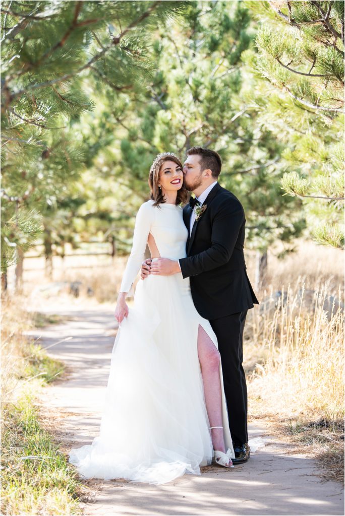 Bride and groom embrace and kiss on a pathway between trees at their colorado wedding venue