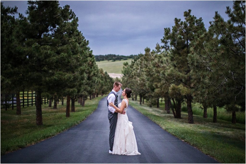 Newly wed couple stands on a black road lined with evergreen trees at their summer wedding in Colorado