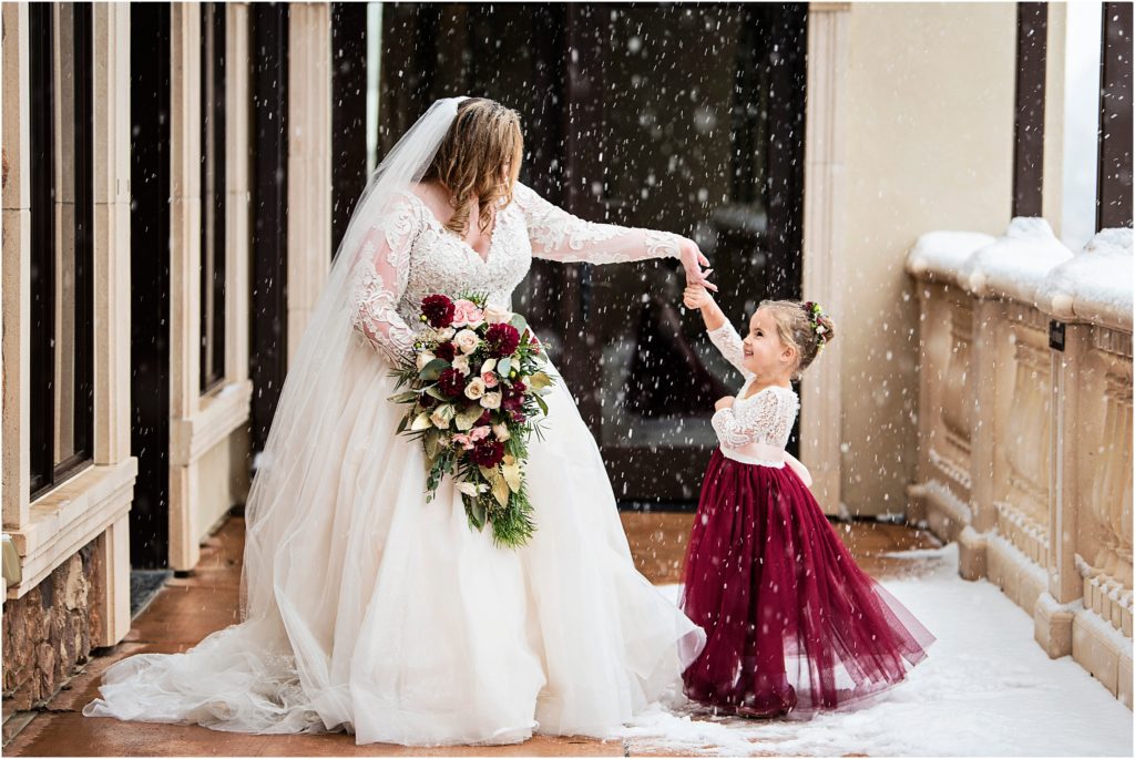 Bride twirls flower girl in the snow holding a bouquet of maroon and pink roses and winter green filler